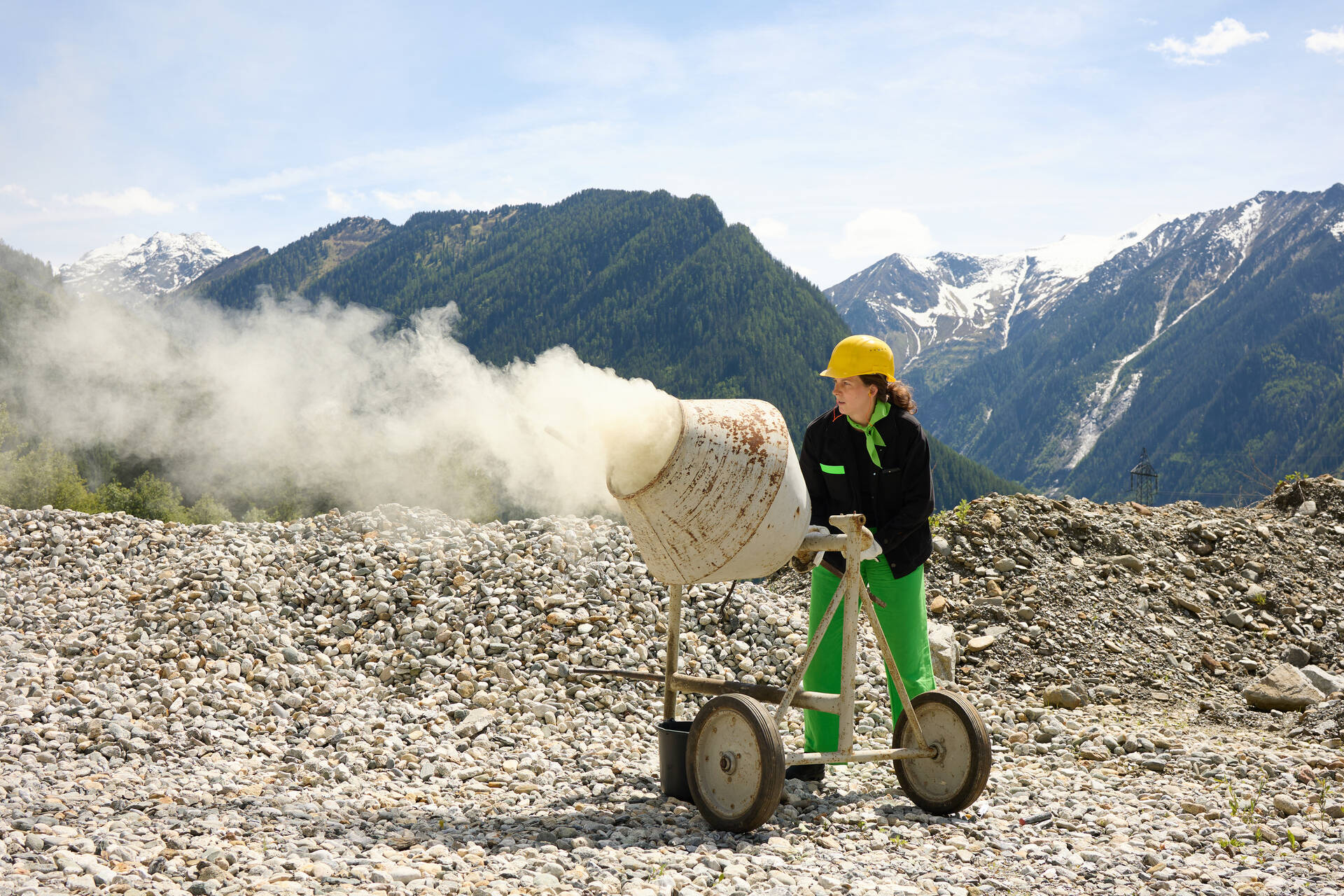 Eine Person steht an einem rauchenden Betonmischer, die Berge im Hintergrund